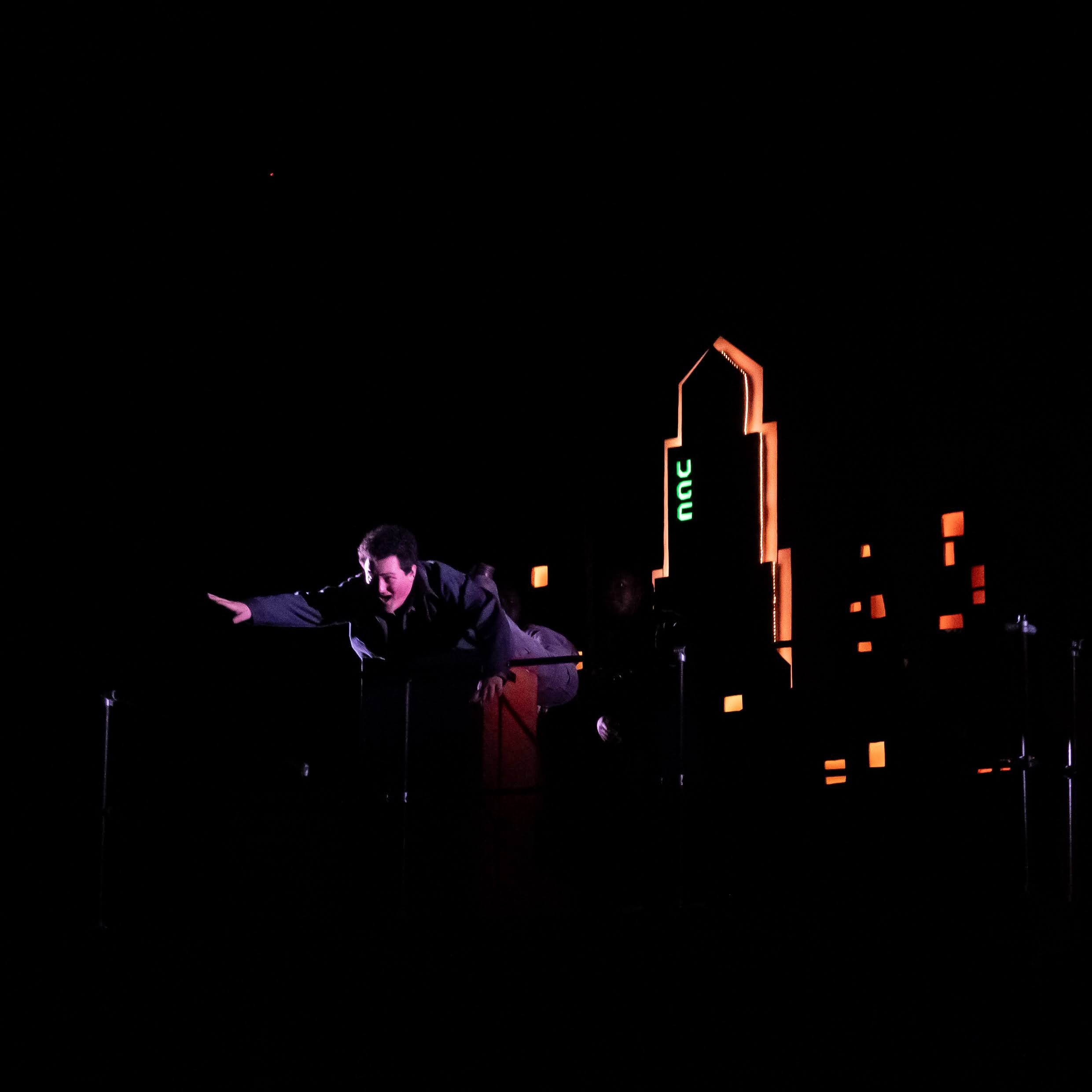 A person in a jumpsuit reaches out as he pretends to fall in front of a lit up, stylized city scape.